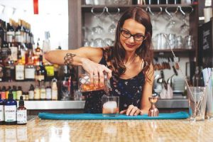 Image Description: Marlene Thorne pours a cocktail from a shaker into a glass, standing behind a clean brightly lit bar,