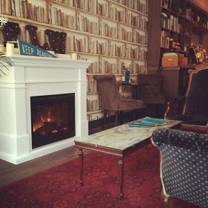 Image Description: Photo of the inside of Famous Last Words. The walls are lined with bookshelf wallpaper, a table sits in front of a fireplace, and the edge of a dark coloured couch can be seen.